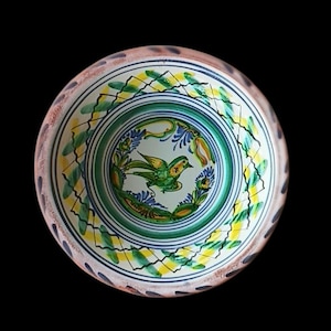 May include: A ceramic bowl with a hand-painted design featuring a green bird in the center, surrounded by green, yellow, and blue floral patterns. The rim has a speckled, light brown and white finish. The bowl is set against a black background.