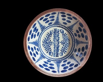 May include: A ceramic bowl with a cream-colored interior and a brown rim. The bowl is decorated with blue floral and geometric patterns. The design includes stylized leaves and dotted accents, creating a folk art aesthetic. The bowl is set against a black background.