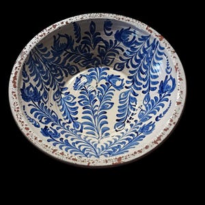 Op de afbeelding: Een ronde keramische kom met een witte binnenkant en een blauw bloemmotief. De rand heeft een verweerde, rustieke uitstraling met bruine en witte accenten. De binnenkant van de kom heeft een gedetailleerd, symmetrisch patroon van blauwe bladeren en bloemen.