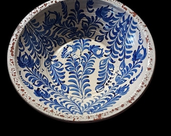 May include: A round, ceramic bowl with a white interior and a blue floral design. The rim has a distressed, rustic look with brown and white accents. The bowl's interior features a detailed, symmetrical pattern of blue leaves and flowers.