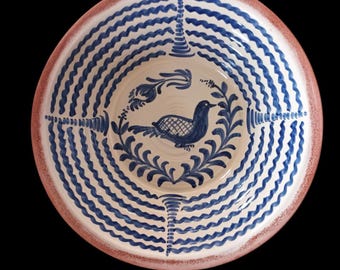 May include: A round ceramic bowl with a hand-painted design. The bowl features a blue bird surrounded by a leafy wreath in the center. The rim has a red-brown border, and the interior has concentric blue wave patterns.
