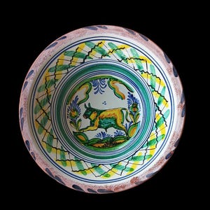 May include: A hand-painted ceramic plate with a bull illustration. The plate features a green, yellow, and blue bull in the center, surrounded by green and blue floral designs. The rim has a decorative pattern with blue and green accents.
