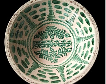 May include: A round, off-white ceramic bowl with a hand-painted green design. The bowl's interior features a central floral motif and a series of green brushstrokes and patterns around the rim. The rim has a textured, brown border.