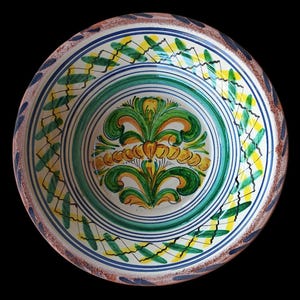 May include: A hand-painted ceramic bowl with a floral design. The bowl features a central motif of green and yellow stylized flowers, surrounded by concentric rings of green, blue, and white. The rim has a brown border with blue and yellow accents.
