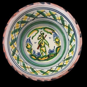 May include: A decorative ceramic bowl with a hand-painted design. The bowl features a central image of a person holding a staff, surrounded by floral and leaf motifs. The rim has a repeating pattern of blue and black dots.