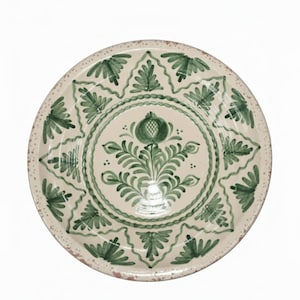 May include: A round, off-white ceramic plate with a green floral design. The plate features a central floral motif and a repeating leaf pattern around the rim. The rim has a distressed, speckled appearance.