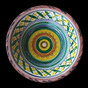 May include: A hand-painted ceramic bowl with a vibrant, concentric design. The center features yellow, brown, and green rings, surrounded by blue and green bands. The rim has a brown border with dark leaf-like patterns. A decorative piece.
