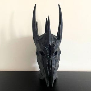 May include: A black 3D printed mask with a pointed crown and detailed facial features. The mask is made of a smooth, matte plastic.