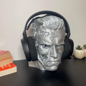 May include: A silver-coloured bust of a man with a robotic face, wearing black headphones. The bust is used as a headphone stand.