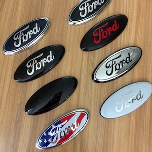 May include: A collection of oval-shaped Ford emblems in various colors, including black, red, silver, and a patriotic design with red and blue stripes and stars. The Ford logo is prominently displayed in a cursive font on each emblem.