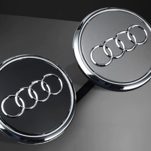 May include: Two round car emblems with a silver chrome border and a black or gray background. The emblems feature the Audi logo, which is four interlocking silver rings.