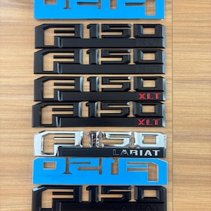 May include: A collection of Ford F-150 truck emblem badges. The badges are in various colors, including chrome, black, and blue. Some badges read "XLT" and "LARIAT". The badges are arranged vertically on a wooden surface.