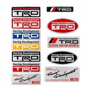 May include: A collection of twelve rectangular and oval metal emblems featuring the TRD Racing Development logo in various color combinations including black, red, blue, yellow, and silver, with some displaying Toyota Motor Sports text.