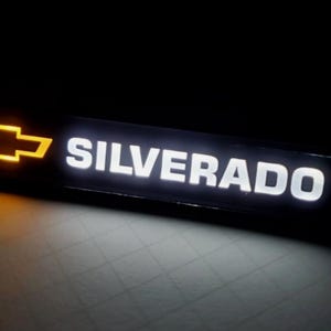 May include: Illuminated black rectangular sign with the word "SILVERADO" in bright white letters. The Chevrolet logo, a gold bowtie, is on the left. The sign is lit against a dark background.