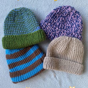 May include: Four knitted beanies in various colors. One is green and blue, another is blue and brown striped, a third is purple and pink, and the last is tan. The beanies are folded at the brim.