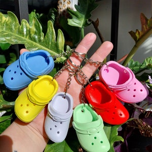 May include: A set of seven colourful mini clog keychains. The keychains are made of a soft, rubbery material and feature a variety of colours, including blue, yellow, pink, red, and green. Each keychain has a small metal ring for attaching to keys or bags.