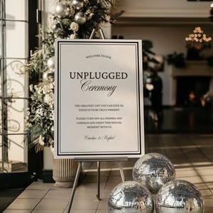 May include: A white sign on an easel with the text "Welcome to our Unplugged Ceremony." The sign is framed in black and has additional text below. Three mirrored disco balls are in front of the sign.