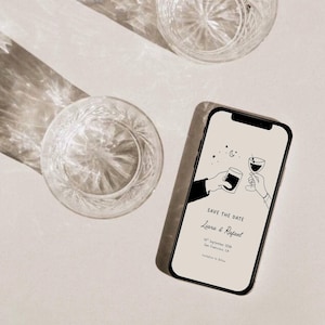 K&ouml;nnte beinhalten: Ein Smartphone zeigt eine Hochzeitseinladung mit dem Text &bdquo;Save the Date&ldquo; und den Namen &bdquo;Liana & Rafael&ldquo;. Zwei Kristallgl&auml;ser sind sichtbar, zusammen mit einem Schatten. Die Einladung zeigt eine Strichzeichnung von zwei H&auml;nden, die ansto&szlig;en.