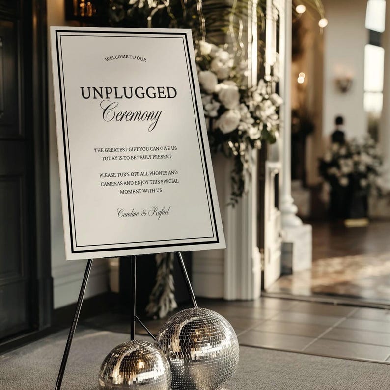 May include: A white sign on a black tripod stand with the text "UNPLUGGED Ceremony". The sign also includes the text "WELCOME TO OUR" and "THE GREATEST GIFT YOU CAN GIVE US TODAY IS TO BE TRULY PRESENT". Two silver disco balls are in front of the sign.