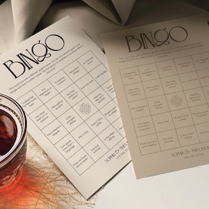 Può includere: Due cartelle di Bingo beige con testo nero, con una griglia di richieste da compilare per gli ospiti. Le cartelle includono i nomi "SOPHIA & THEO" e la data "09.08.2026". Un bicchiere di liquido rosso è visibile nell'angolo.