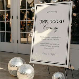 May include: A white sign with black text that reads "WELCOME TO OUR UNPLUGGED Ceremony." The sign is on a wooden easel, with two silver disco balls in front. The sign also includes the text "Caroline & Rafael."