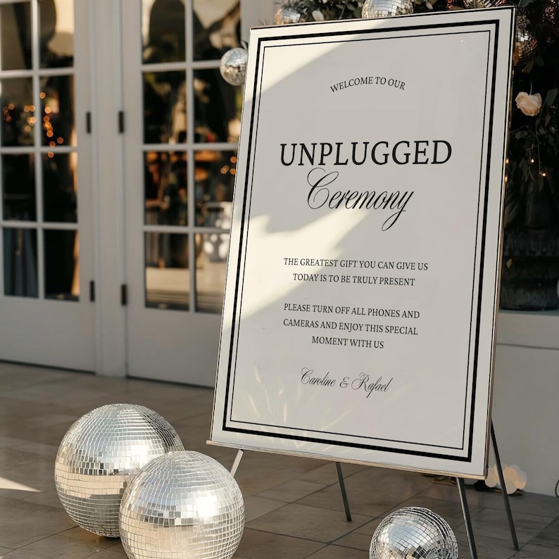 May include: A white sign with black text that reads "WELCOME TO OUR UNPLUGGED Ceremony." The sign is on a wooden easel, with two silver disco balls in front. The sign also includes the text "Caroline & Rafael."