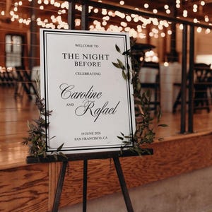 May include: A welcome sign on a black easel, announcing "The Night Before" celebrating Caroline and Rafael, with the date June 19, 2026, in San Francisco, CA. The sign is framed in black and decorated with greenery.