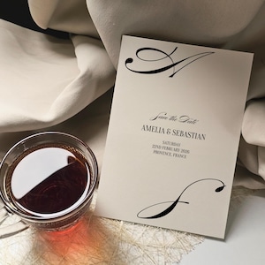 May include: A cream-colored "Save the Date" card with elegant black calligraphy. The card features the names Amelia & Sebastian, the date, and location. A glass of tea sits beside the card, adding a touch of sophistication.
