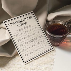 Peut inclure: Une carte de jeu de Bingo beige avec du texte noir qui dit "Find the Guest Bingo". La carte a une grille de cases avec des indices. Une tasse &agrave; th&eacute; en verre transparent remplie de th&eacute; se trouve &agrave; droite de la carte.