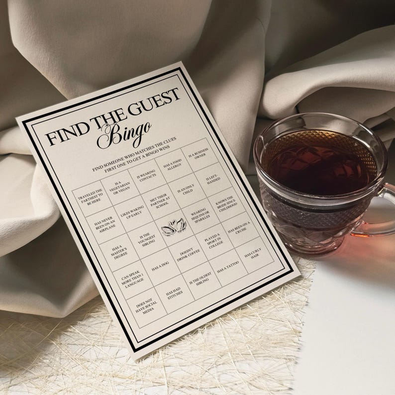 Peut inclure: Une carte de jeu de Bingo beige avec du texte noir qui dit "Find the Guest Bingo". La carte a une grille de cases avec des indices. Une tasse &agrave; th&eacute; en verre transparent remplie de th&eacute; se trouve &agrave; droite de la carte.