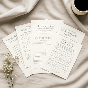 Pu&ograve; includere: Una collezione di biglietti di giochi per matrimoni bianchi con testo nero, tra cui "He Said", "Wedding Word Search", "I Love You Around the Wo", "Guest Who?", "Wedding Photo Scavenger Hunt" e "Find the Guest Bingo". Una tazza di caff&egrave; &egrave; sullo sfondo.