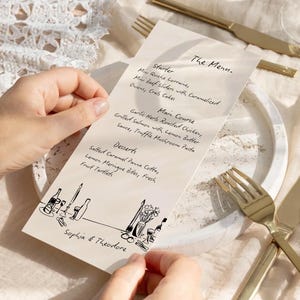May include: A menu card with handwritten text, held by hands, featuring a list of dishes. The card is cream-colored with black illustrations of wine bottles and glasses. Gold-colored cutlery and a white plate are also visible.