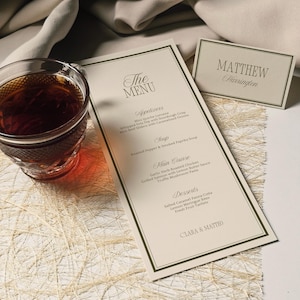 May include: A formal menu and place card set. The menu features a cream-colored background with dark green borders and text. The menu includes appetizers, soup, main course, and desserts. A glass of tea sits to the left of the menu.
