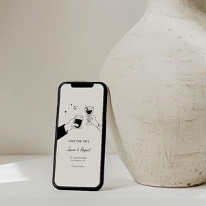 K&ouml;nnte beinhalten: Ein Smartphone zeigt eine minimalistische Hochzeitsank&uuml;ndigung. Der Bildschirm zeigt eine Strichzeichnung von zwei H&auml;nden, die mit Getr&auml;nken ansto&szlig;en, zusammen mit dem Text "Save the Date" und Namen. Das Telefon ist schwarz und befindet sich in der N&auml;he einer beigen Vase.