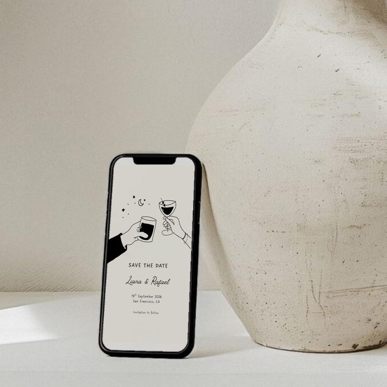 K&ouml;nnte beinhalten: Ein Smartphone zeigt eine minimalistische Hochzeitsank&uuml;ndigung. Der Bildschirm zeigt eine Strichzeichnung von zwei H&auml;nden, die mit Getr&auml;nken ansto&szlig;en, zusammen mit dem Text "Save the Date" und Namen. Das Telefon ist schwarz und befindet sich in der N&auml;he einer beigen Vase.