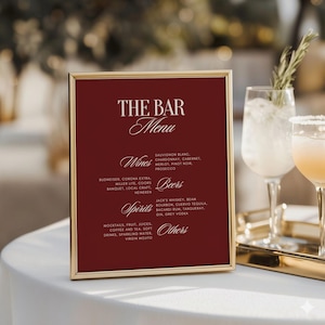Op de afbeelding: Een ingelijste barmenu met een bordeauxrode achtergrond en een gouden rand. Het menu bevat wijnen, bieren, sterke drank en andere drankopties. Op de achtergrond zijn twee cocktails te zien.