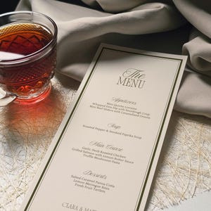 May include: A menu card with a cream background and a dark green border. The menu lists appetizers, soup, main courses, and desserts. A glass of tea sits to the left of the menu, and a cream-colored cloth is in the background.