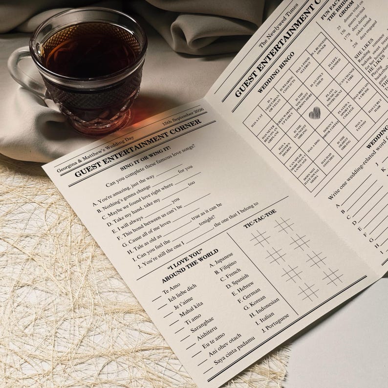 Pu&ograve; includere: Una tazza di t&egrave; di vetro trasparente piena di t&egrave; scuro &egrave; appoggiata su un panno bianco accanto a un libretto di giochi per matrimoni. Il libretto presenta giochi a tema matrimonio, tra cui un gioco di bingo e un gioco di canzoni da completare. Il testo sul libretto recita "Guest Entertainment Corner".