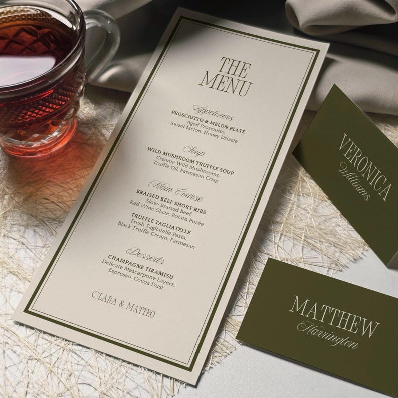 May include: Elegant menu card with a cream background and dark green border, featuring a detailed menu with appetizers, soup, main course, and desserts. Includes two name cards with names. A glass of tea is in the background.