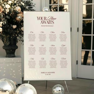 May include: A white wedding seating chart on a metal easel. The chart features a burgundy font with the words "YOUR Place AWAITS" and guest names organized by table number. Disco balls are in the foreground.