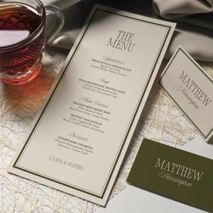 May include: Elegant wedding menu with a cream-colored background and a dark green border. The menu features appetizers, soup, main course, and desserts. A matching name card is also present, along with a glass of tea.