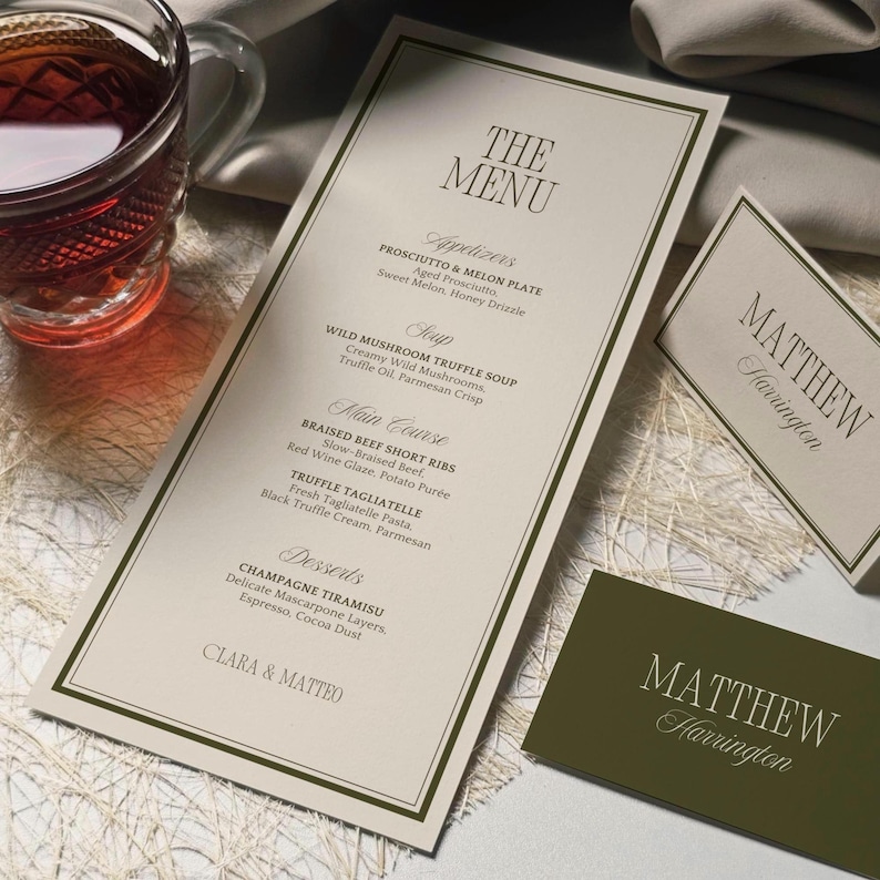 May include: Elegant wedding menu with a cream-colored background and a dark green border. The menu features appetizers, soup, main course, and desserts. A matching name card is also present, along with a glass of tea.