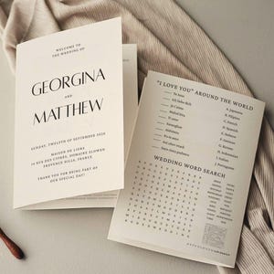 May include: Elegant wedding stationery featuring the names "GEORGINA AND MATTHEW" in a stylish font. Includes a wedding program with details and a word search puzzle with phrases like "I love you" in multiple languages.