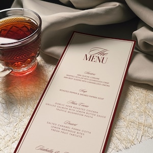 May include: A menu card with a burgundy border and elegant script. The menu lists starters, soup, main courses, and desserts. A glass of tea sits to the left of the menu. The menu is on a white surface with a beige cloth in the background.