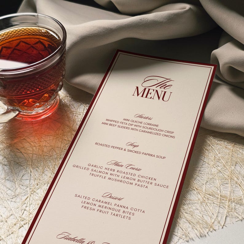 May include: A menu card with a burgundy border and elegant script. The menu lists starters, soup, main courses, and desserts. A glass of tea sits to the left of the menu. The menu is on a white surface with a beige cloth in the background.