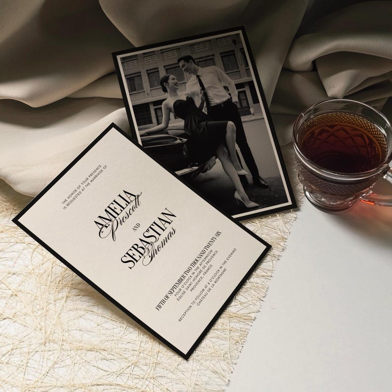 May include: Elegant wedding invitation with black border and text "Amelia Prescott and Sebastian Thomas". A black and white photo of a couple is behind the invitation. A glass teacup with tea is on the right.