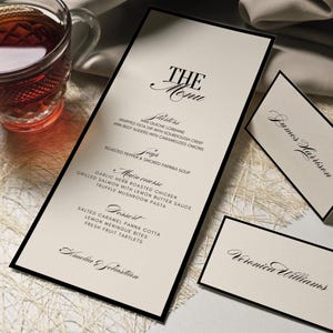 K&ouml;nnte beinhalten: Elegante Men&uuml;karte mit schwarzem Rand und dem Text "THE Menu" in Schreibschrift. Das Men&uuml; listet Vorspeisen, Suppe, Hauptgang und Dessert-Optionen auf. Inklusive Tischkarten mit Namen.