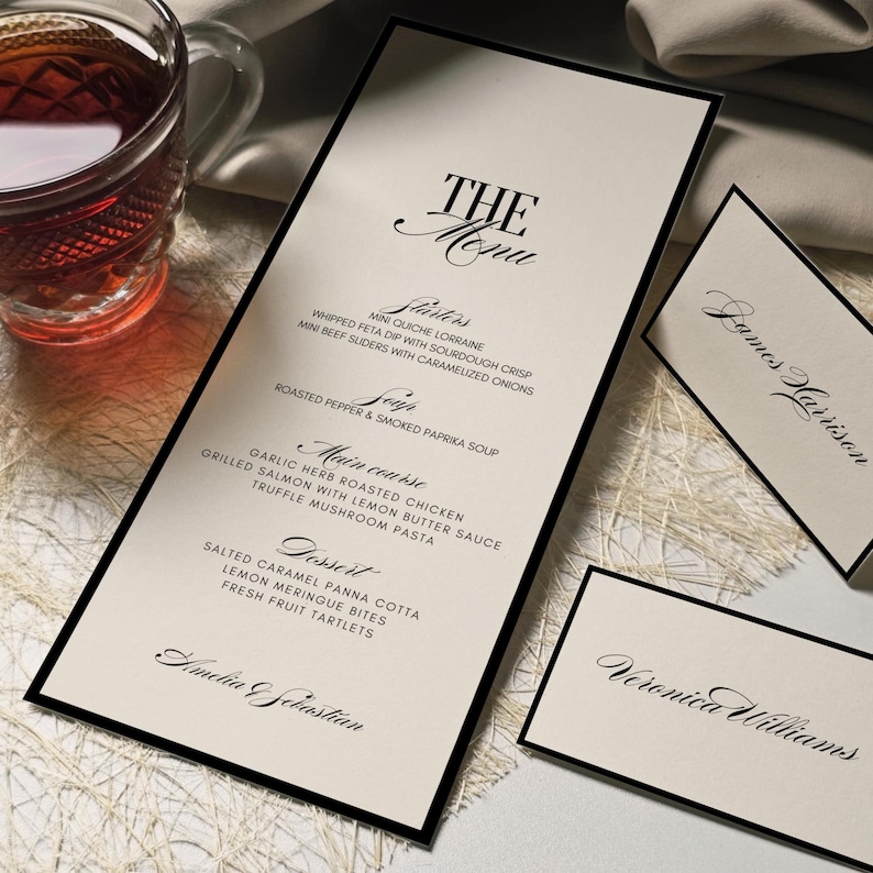 K&ouml;nnte beinhalten: Elegante Men&uuml;karte mit schwarzem Rand und dem Text "THE Menu" in Schreibschrift. Das Men&uuml; listet Vorspeisen, Suppe, Hauptgang und Dessert-Optionen auf. Inklusive Tischkarten mit Namen.