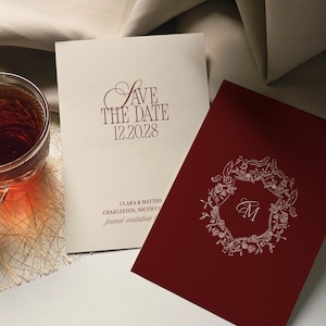 May include: Two wedding invitation cards. One is cream-colored with burgundy text that reads "Save the Date 12.20.28" and the names "Clara & Matteo" and "Charleston, South Carolina". The other is burgundy with a floral wreath and the letter "M".