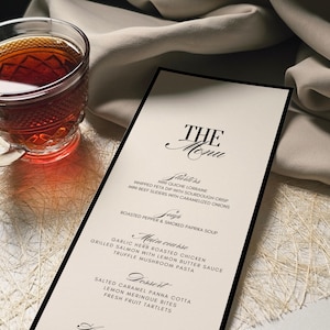 K&ouml;nnte beinhalten: Eine Speisekarte mit schwarzem Rand und dem Titel "THE Menu" in eleganter Schrift. Die Speisekarte listet Vorspeisen, Suppen, Hauptgerichte und Desserts auf. Links vom Men&uuml; steht eine Teetasse aus Glas mit einer dunklen Fl&uuml;ssigkeit.